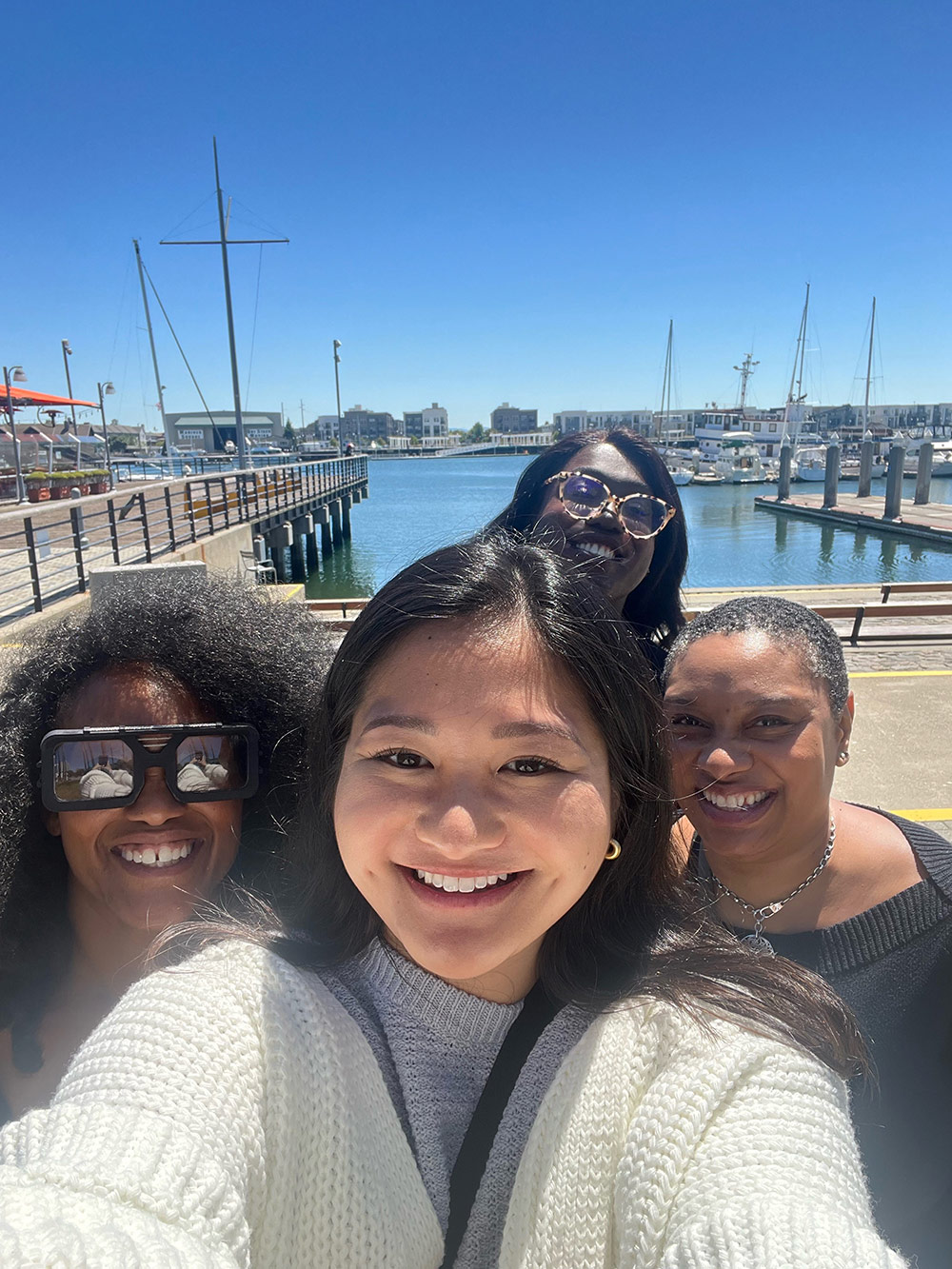 Four members of our Diversity, Equity and Inclusion team stand close together on a pier, with blue water and blue sky behind them